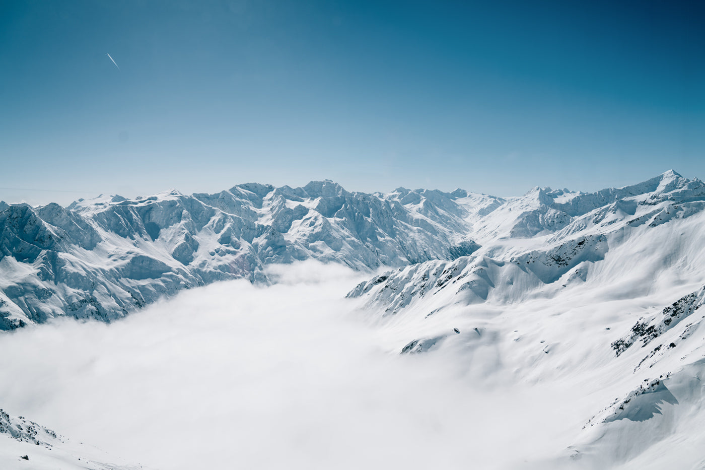 A breathtaking aerial view of snow-covered mountains under a clear blue sky. The rugged peaks rise above a layer of white clouds, creating a serene winter landscape.
