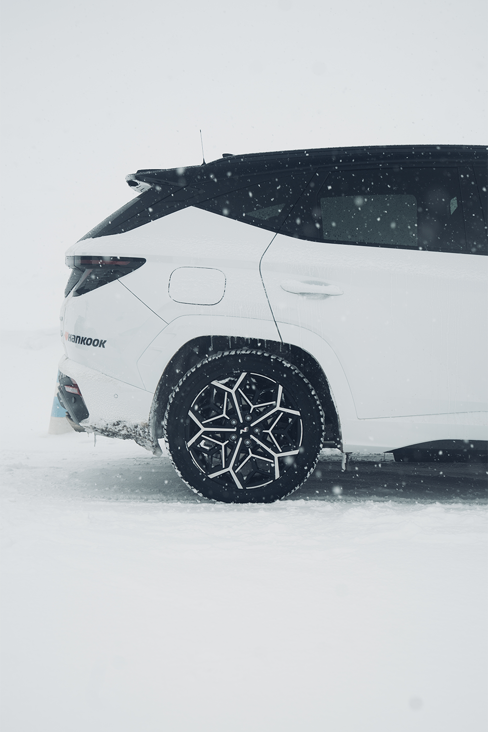 A close-up shot of the rear wheel of a Hyundai Tucson N Line in a snowy setting, showcasing its unique snowflake-patterned alloy wheel design, covered in light snowfall.
