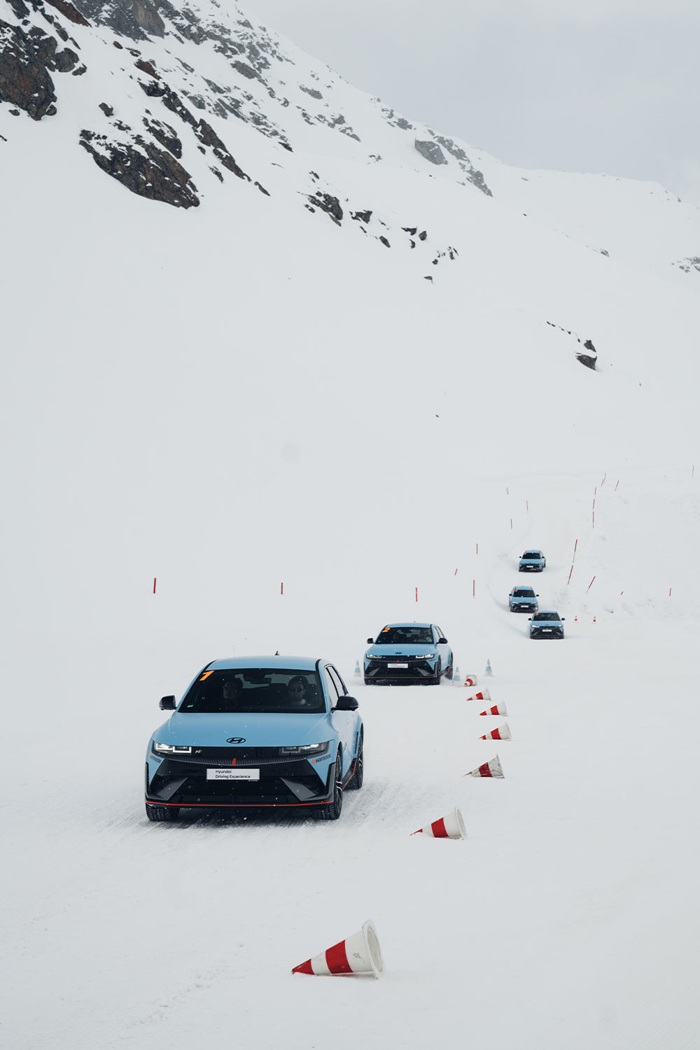 A convoy of Hyundai IONIQ 5 N vehicles driving on a snow-covered mountain track, surrounded by scattered traffic cones, showcasing their capabilities in extreme winter conditions.
