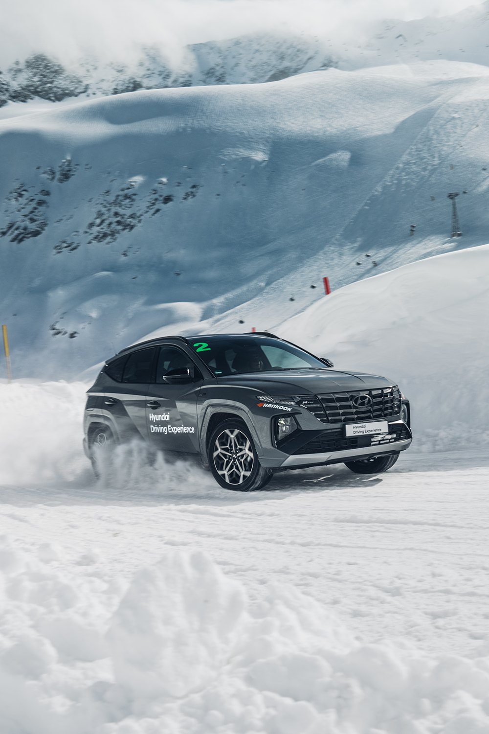 A Hyundai Tucson N Line SUV participating in the off-road Hyundai Driving Experience, driving through snowy terrain with a snowy mountain backdrop.
