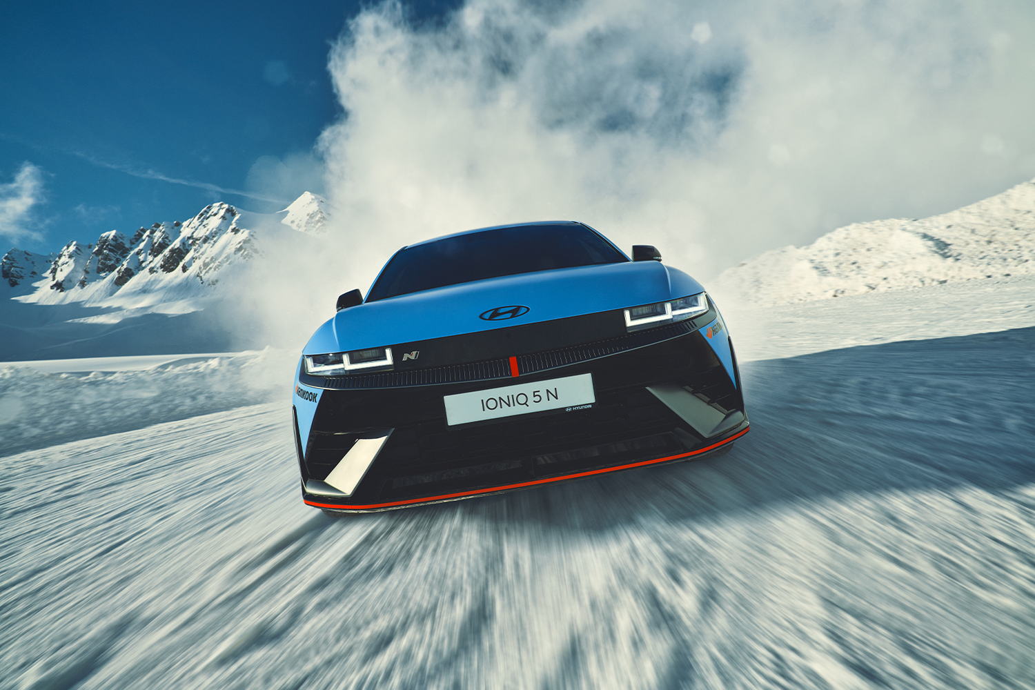 A Hyundai IONIQ 5 N in vibrant blue speeding across a snowy terrain under a clear blue sky, leaving a trail of snow dust, with snow-covered mountains in the background.
