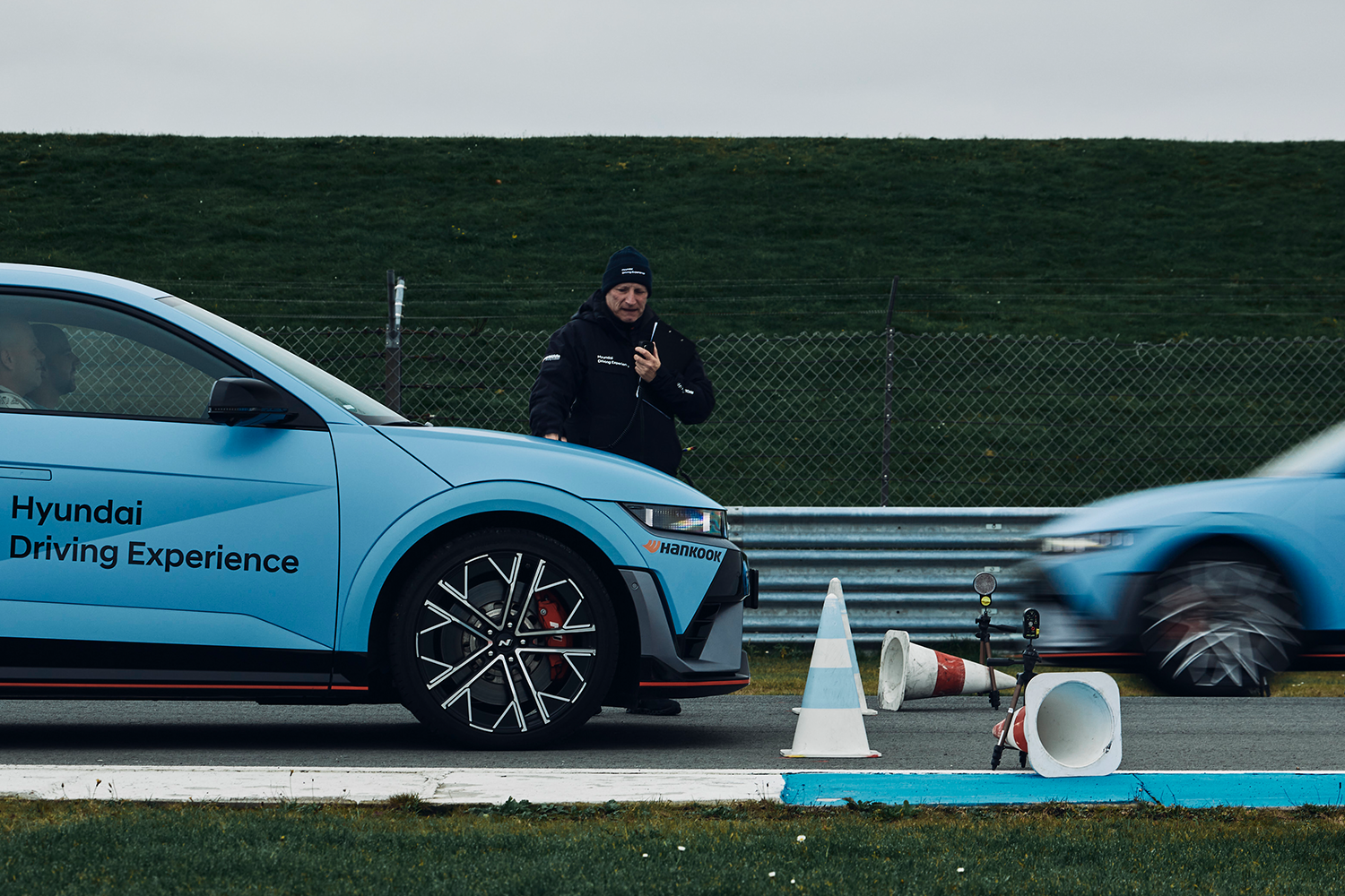 A Hyundai IONIQ 5 N at a Hyundai Driving Experience event, with an instructor using a radio and another IONIQ 5 N speeding past in the background.
