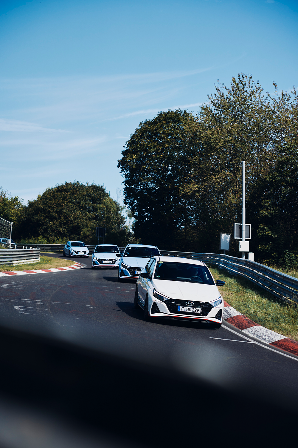 A convoy of Hyundai i20 N cars navigating a turn on a racetrack, surrounded by lush greenery under a clear blue sky.
