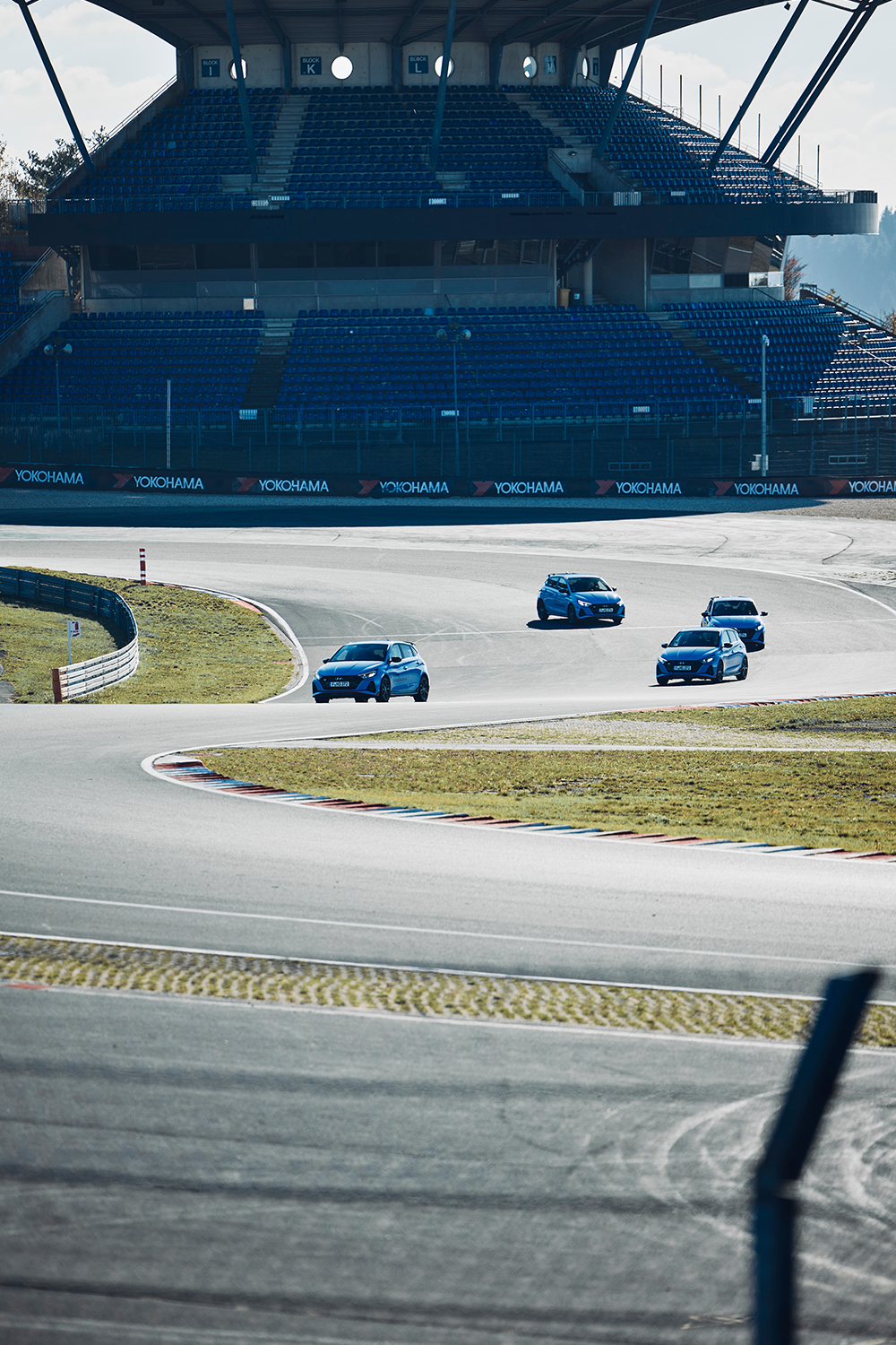 Four Hyundai i20 N cars navigating a turn on a racetrack, with an empty grandstand in the background.
