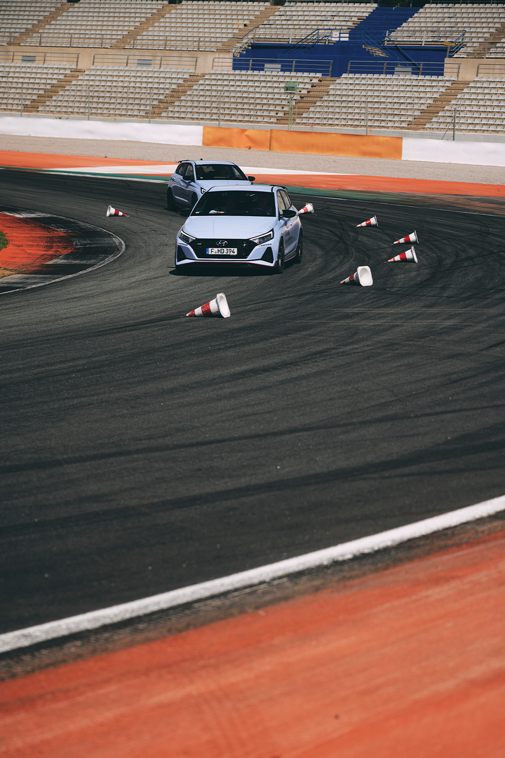 Two Hyundai i20 N cars navigating a sharp turn on a racetrack, knocking over cones, with an empty grandstand in the background.
