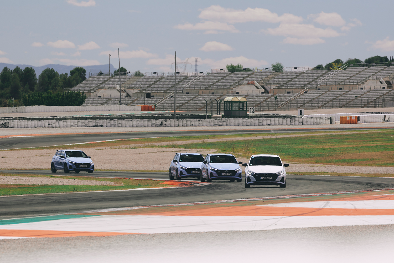 Four Hyundai i20 N cars driving in formation on a racetrack, with an empty grandstand and a clear sky in the background.
