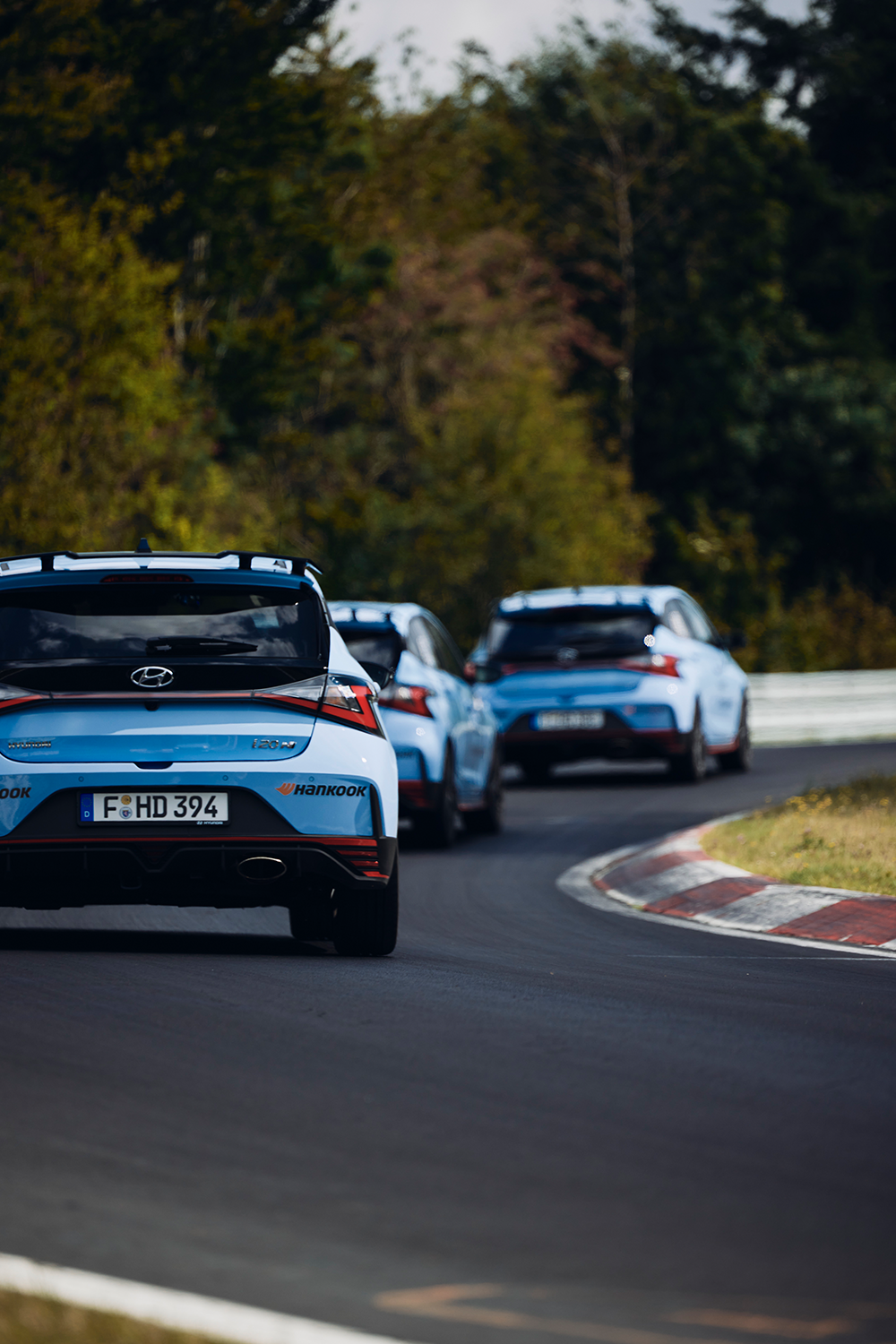 A group of Hyundai i20 N cars in Performance Blue driving in formation on a racetrack, surrounded by lush greenery.
