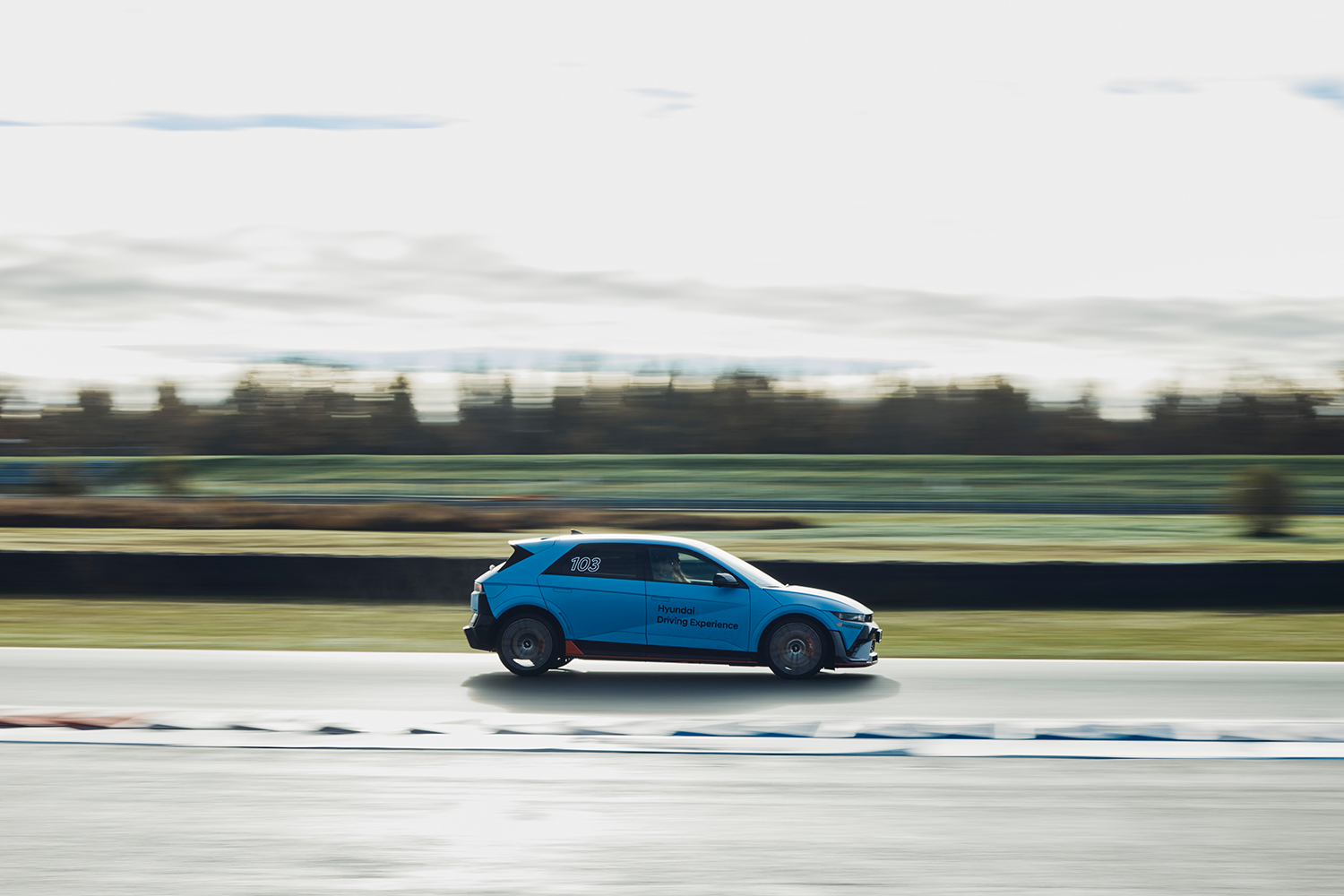 A light blue Hyundai IONIQ 5 N with "Hyundai Driving Experience" branding speeds along a track in motion blur, emphasizing its dynamic performance against a backdrop of blurred greenery and overcast sky.
