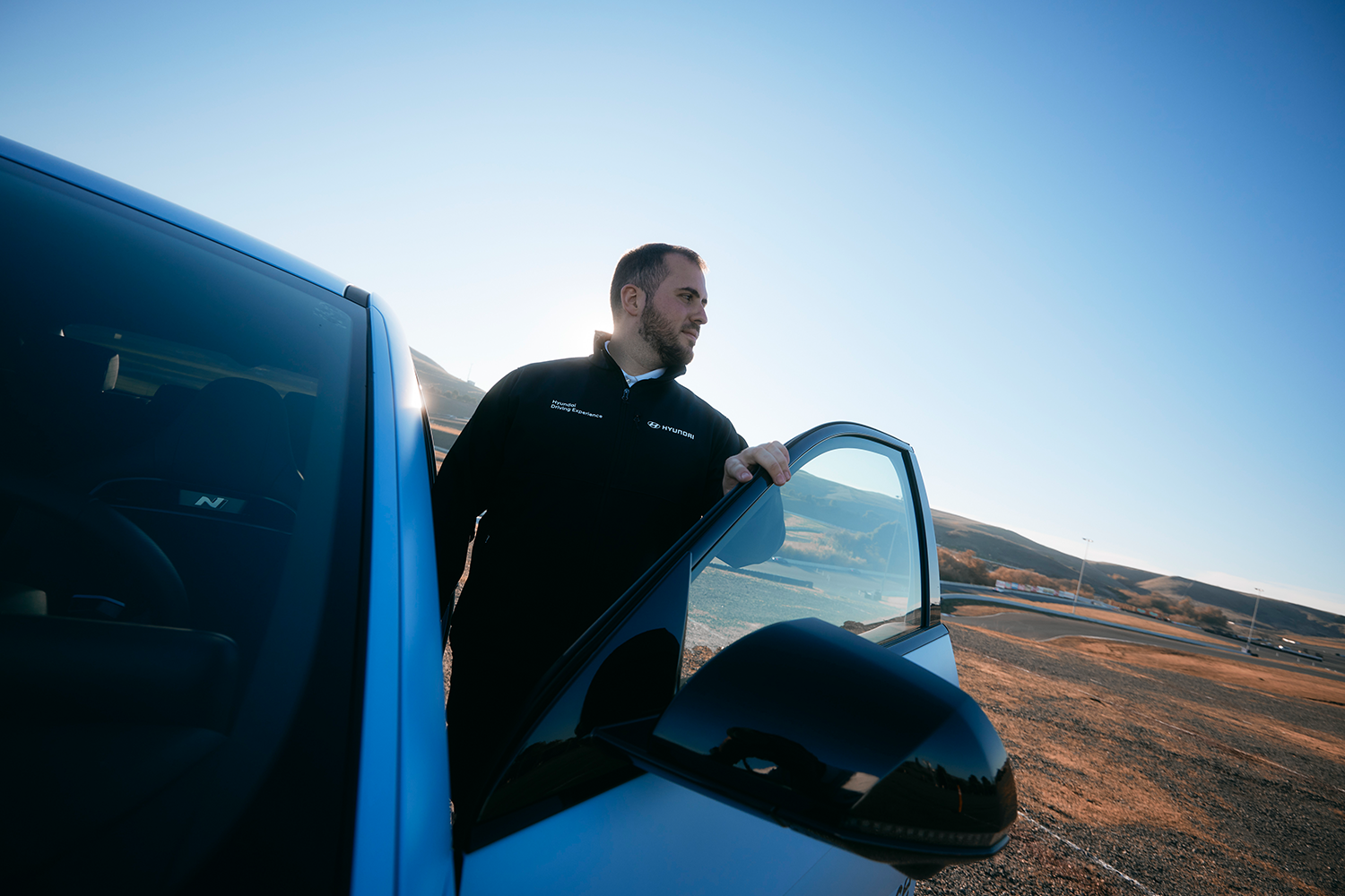 Ricardo stands next to an open door of the Hyundai IONIQ 5 N under a bright blue sky, with a racetrack and hilly landscape in the background.
