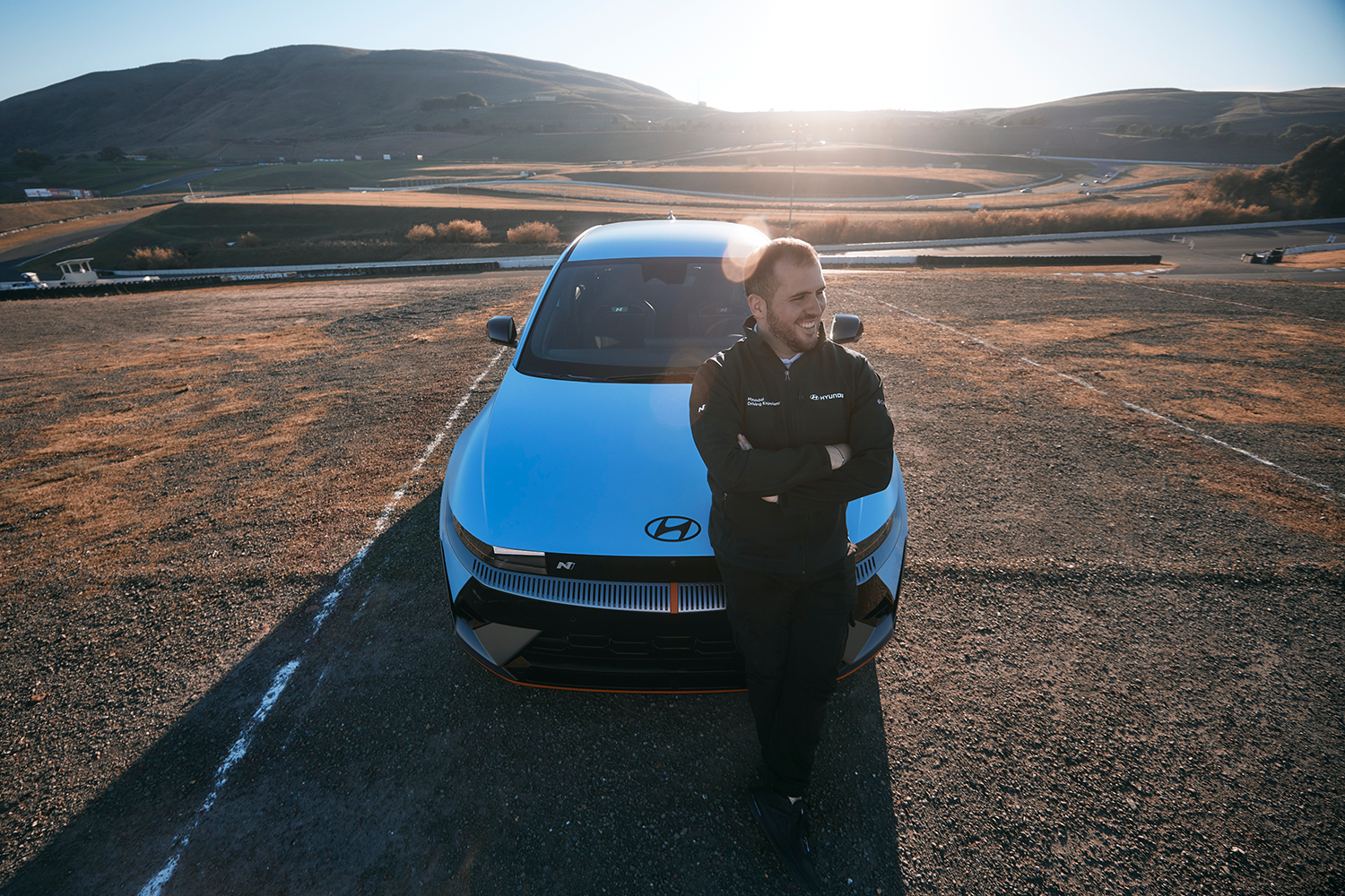 A person stands confidently with arms crossed in front of a Hyundai IONIQ 5 N on a track, with a backdrop of rolling hills and a sunlit circuit in the distance.
