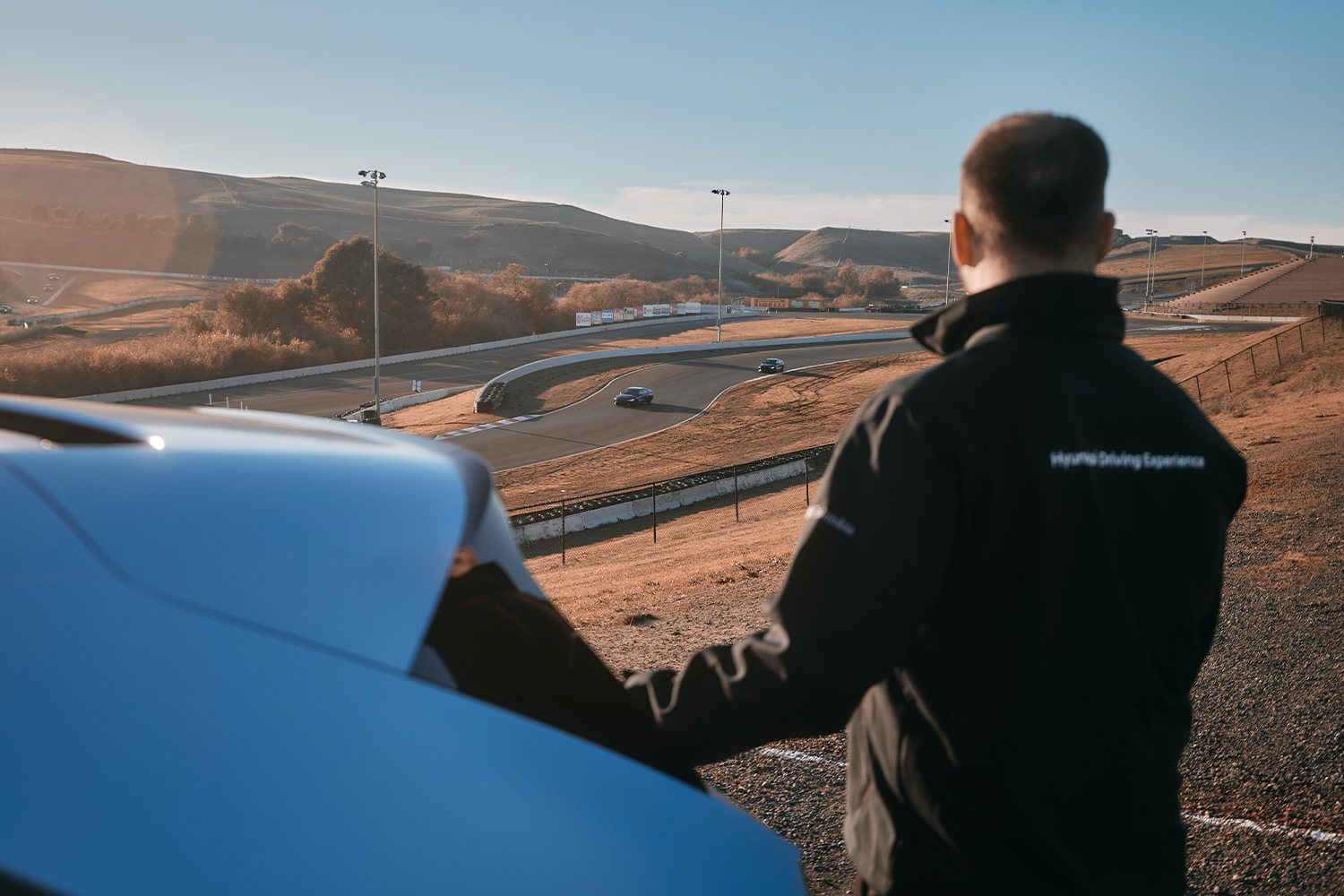 A person wearing a Hyundai Driving Experience jacket stands near a blue Hyundai IONIQ 5 N, overlooking a sunny racetrack with winding curves in the distance. The scene is set in a golden landscape under a clear blue sky.
