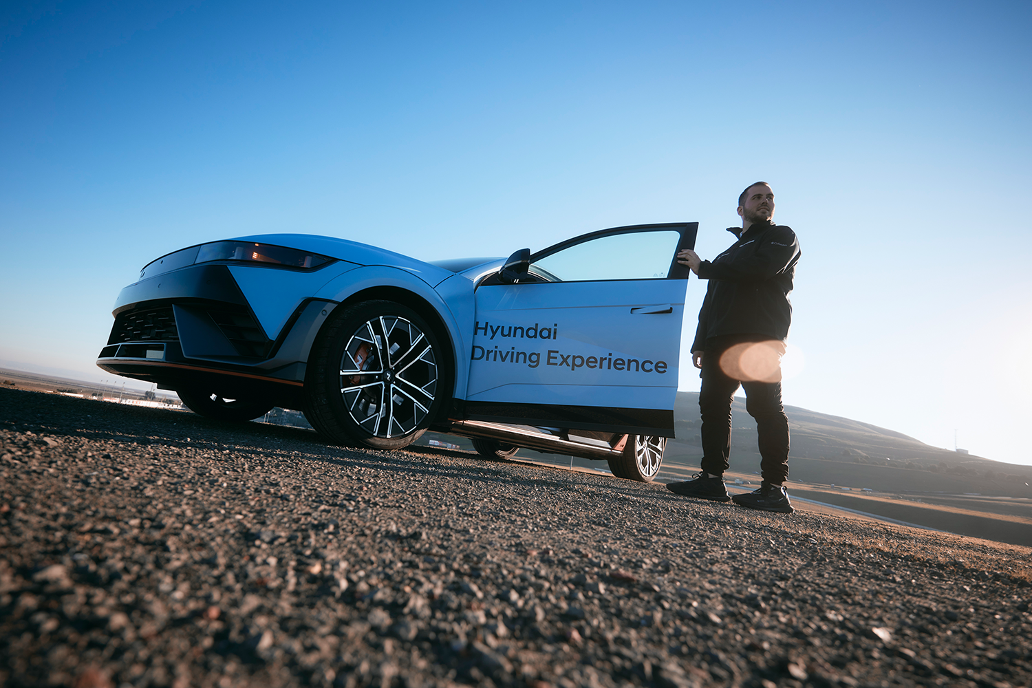 A Hyundai IONIQ 5 N in the "Hyundai Driving Experience" livery parked on a gravel surface during sunrise, with a man standing by the open driver's door looking into the distance. The clear blue sky and distant hills form the backdrop.
