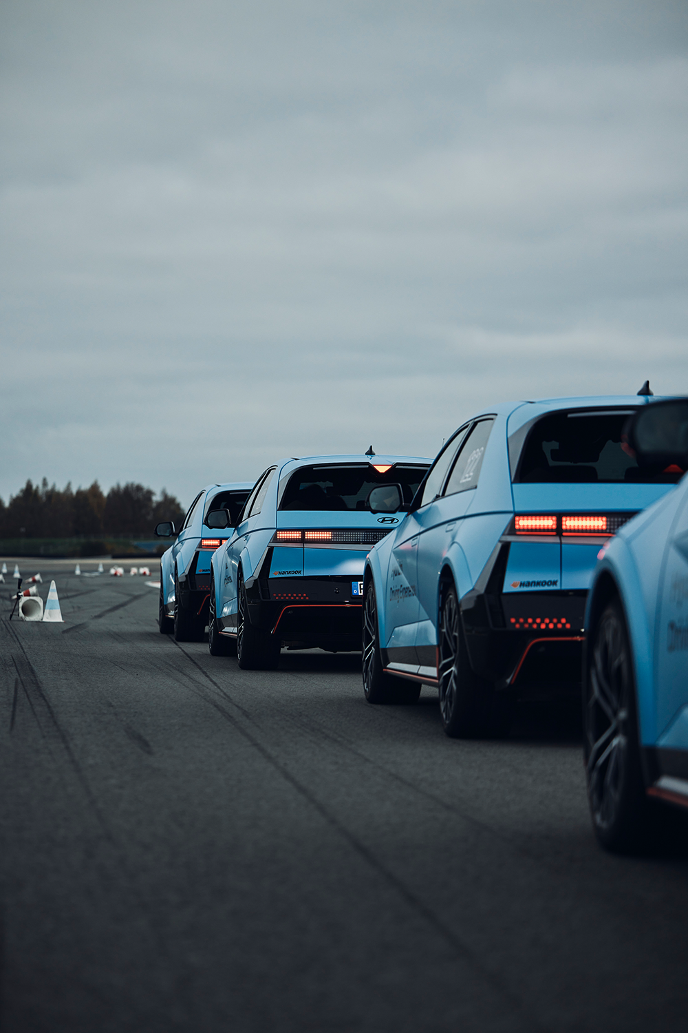 A lineup of Hyundai IONIQ 5 N vehicles seen from the rear, driving in formation on a track during the Hyundai Driving Experience event, with cones marking the course on an overcast day.
