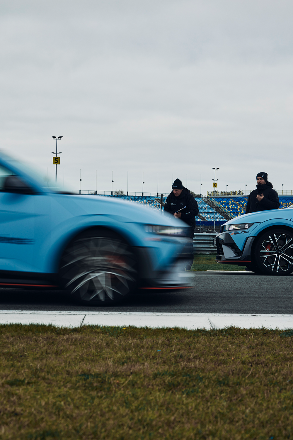 A Hyundai IONIQ 5 N car speeding on a racetrack, with blurred motion indicating high speed, while two individuals in black Hyundai Driving Experience jackets stand near another IONIQ 5 N in the background under an overcast sky.
