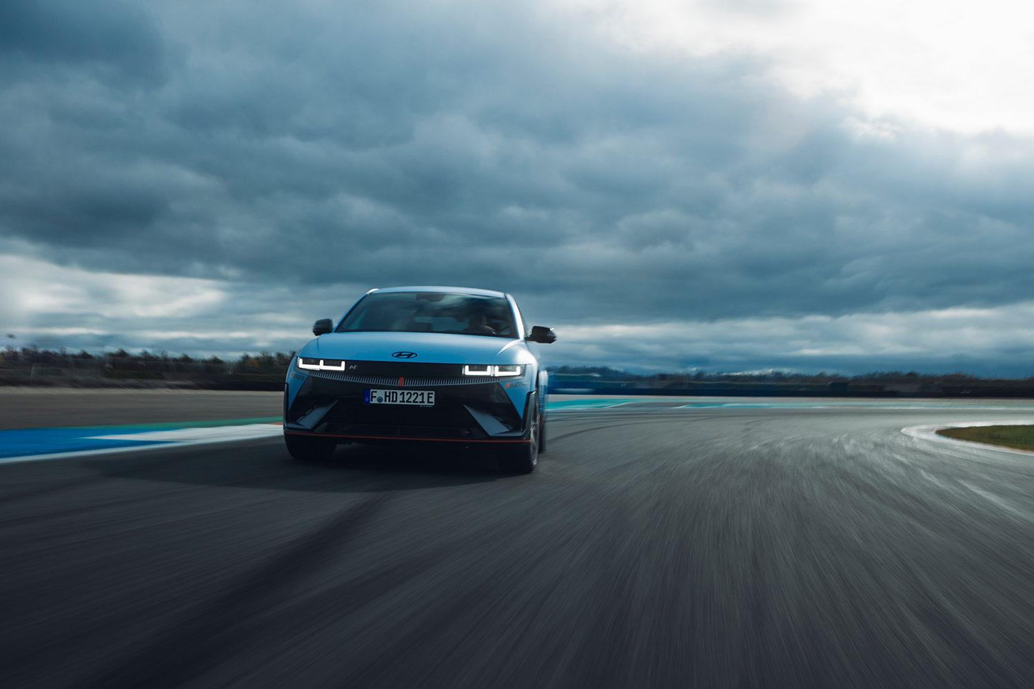 The Hyundai IONIQ 5 N speeds down a racing track under cloudy skies, showcasing its performance capabilities on a sharp curve. The vehicle's distinctive headlights and aerodynamic design are accentuated by the dramatic backdrop.
