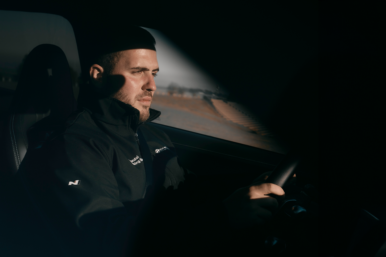 A man focused on driving, sitting in a Hyundai vehicle during a driving experience. The interior is dimly lit, highlighting his concentration as he grips the steering wheel against a backdrop of sunlit landscapes.
