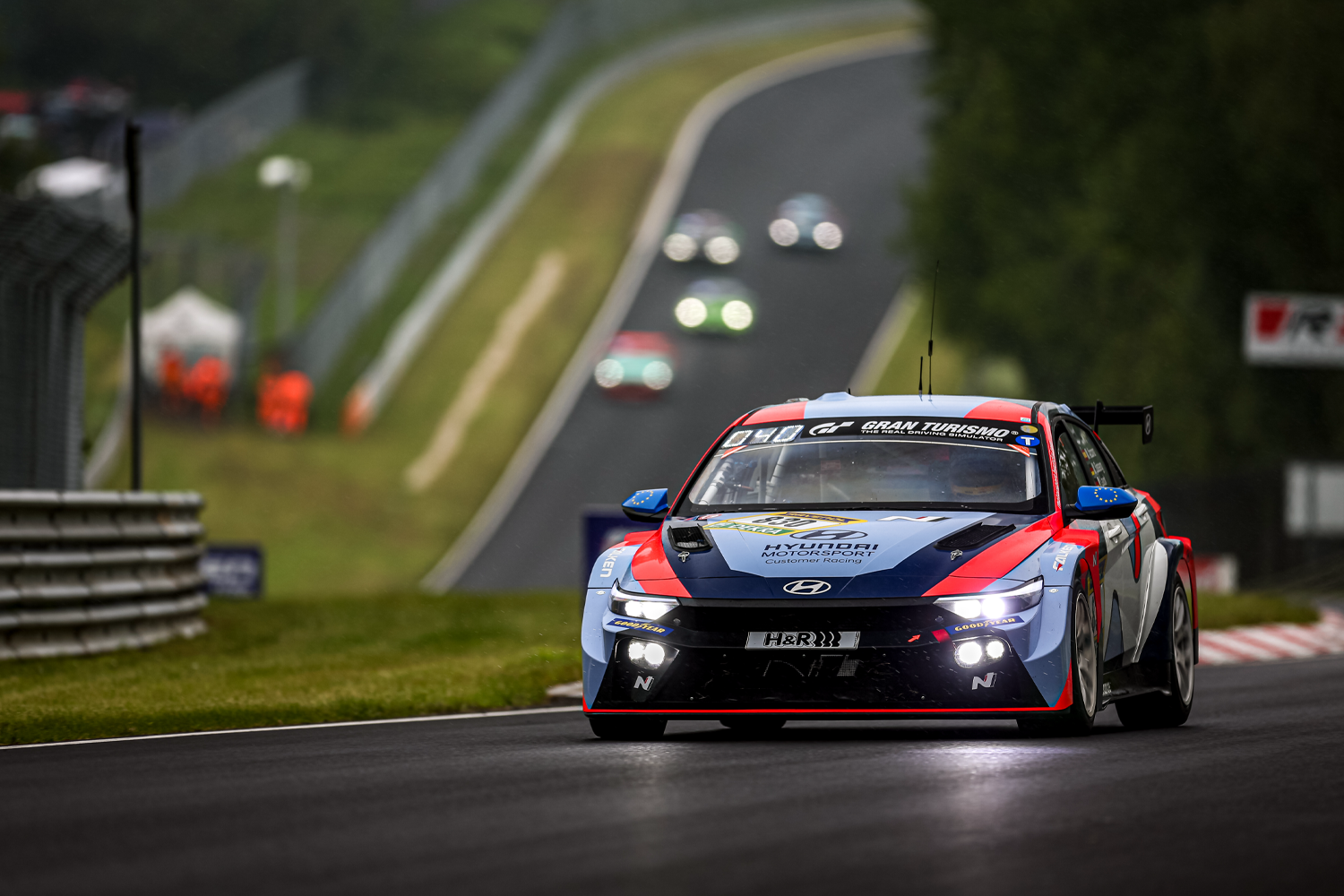 Hyundai Motorsport racing car driving on the Nürburgring track, showcasing its bold livery and performance under competitive conditions with blurred background elements.
