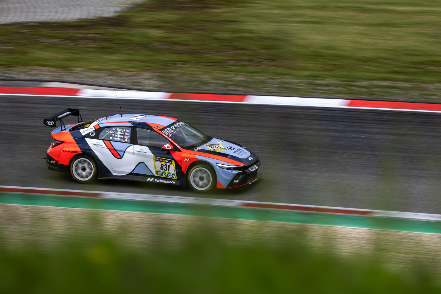 Hyundai N Performance race car speeding through a corner on a wet racetrack, displaying its striking livery and aerodynamic design with motion blur emphasizing its speed.
