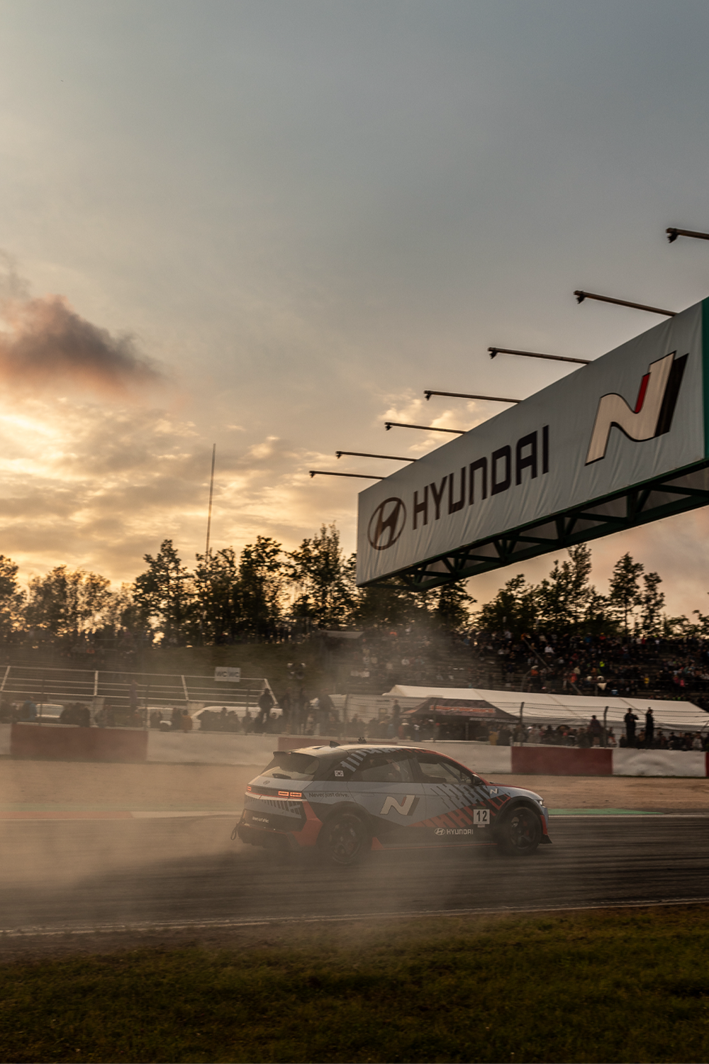 Hyundai N racing car drifting on a track at sunset, with a Hyundai N banner overhead and a smoky atmosphere as spectators watch from the stands.
