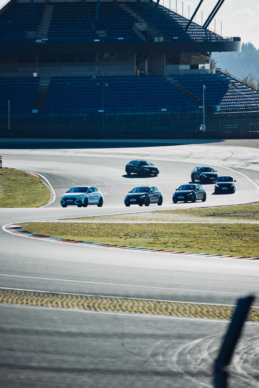 A group of Hyundai vehicles navigating a turn on a racetrack, with a large grandstand in the background, showcasing the Hyundai Driving Experience in action.
