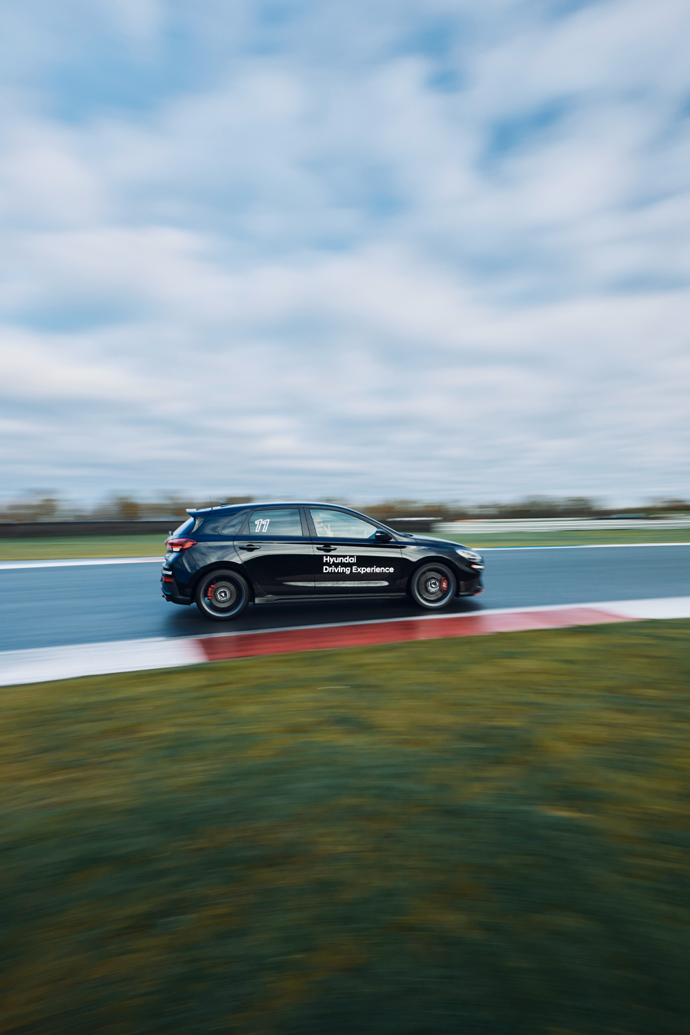Side view of a black Hyundai Driving Experience car driving on a wet racetrack, captured with motion blur, highlighting its dynamic performance and sporty design.
