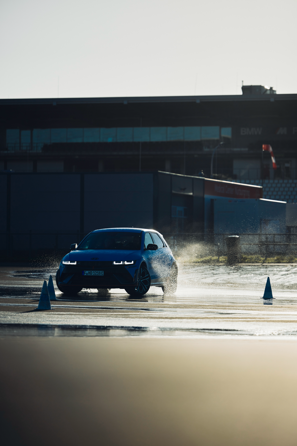 Hyundai IONIQ 5 N performing a wet handling test on a track, splashing water as it navigates through cones, with a building and clear sky in the background.
