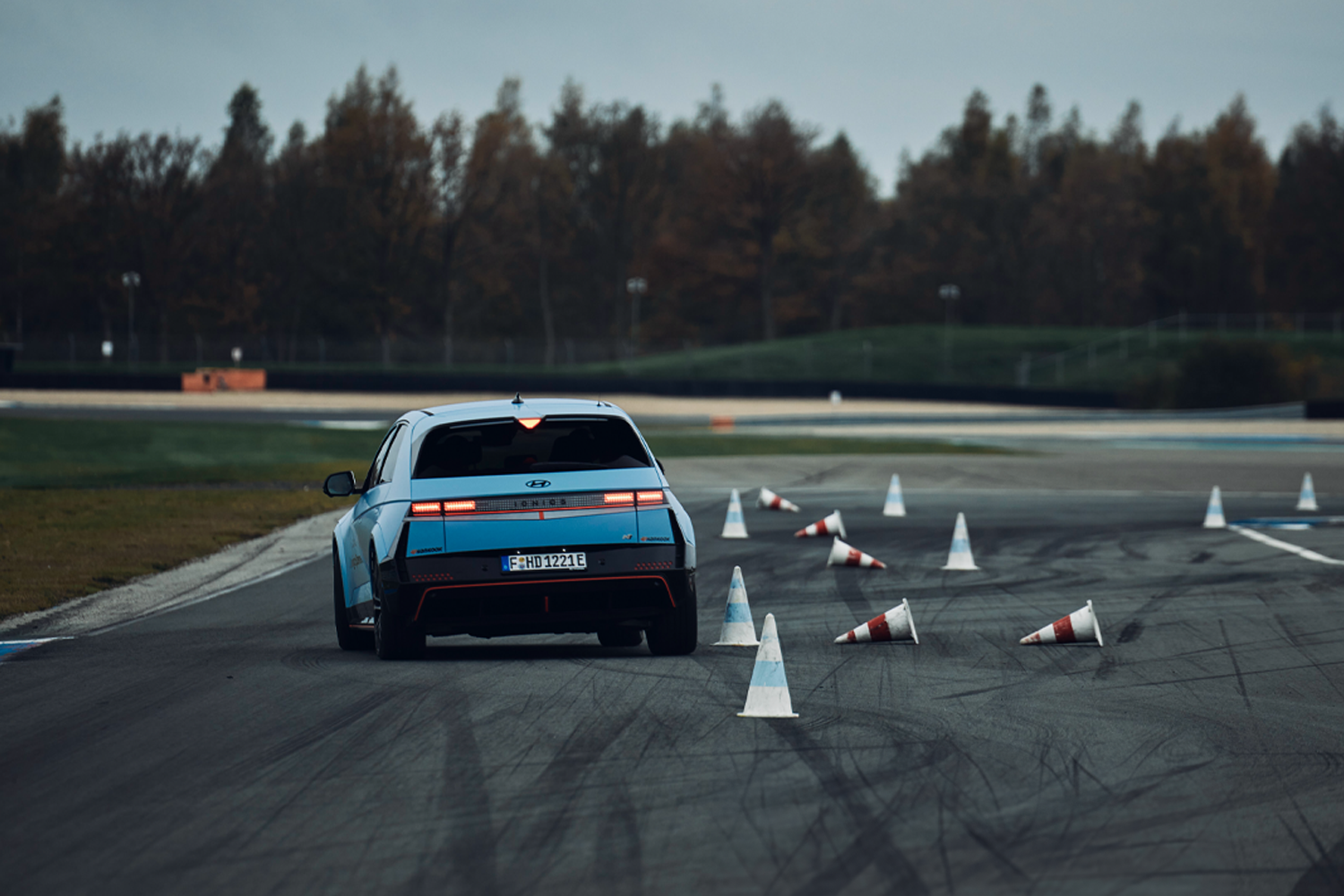 Rear view of the Hyundai IONIQ 5 N navigating through a slalom course marked with cones on a racetrack, with a forested background and tire marks on the asphalt.

