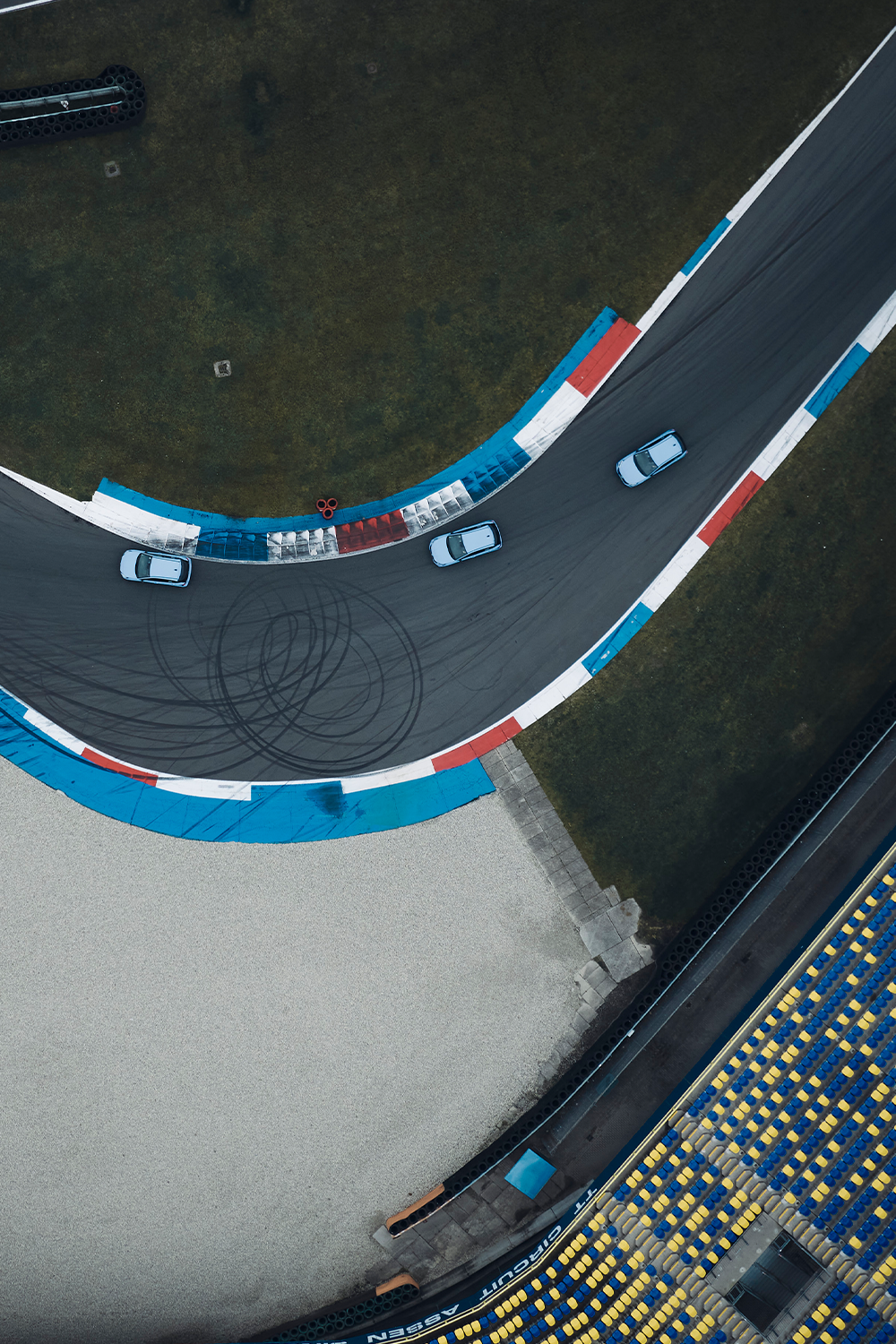 Aerial view of three Hyundai vehicles navigating a sharp turn on a racetrack, with tire marks visible on the asphalt and blue, red, and white curbing lining the track, surrounded by green grass and spectator seating.
