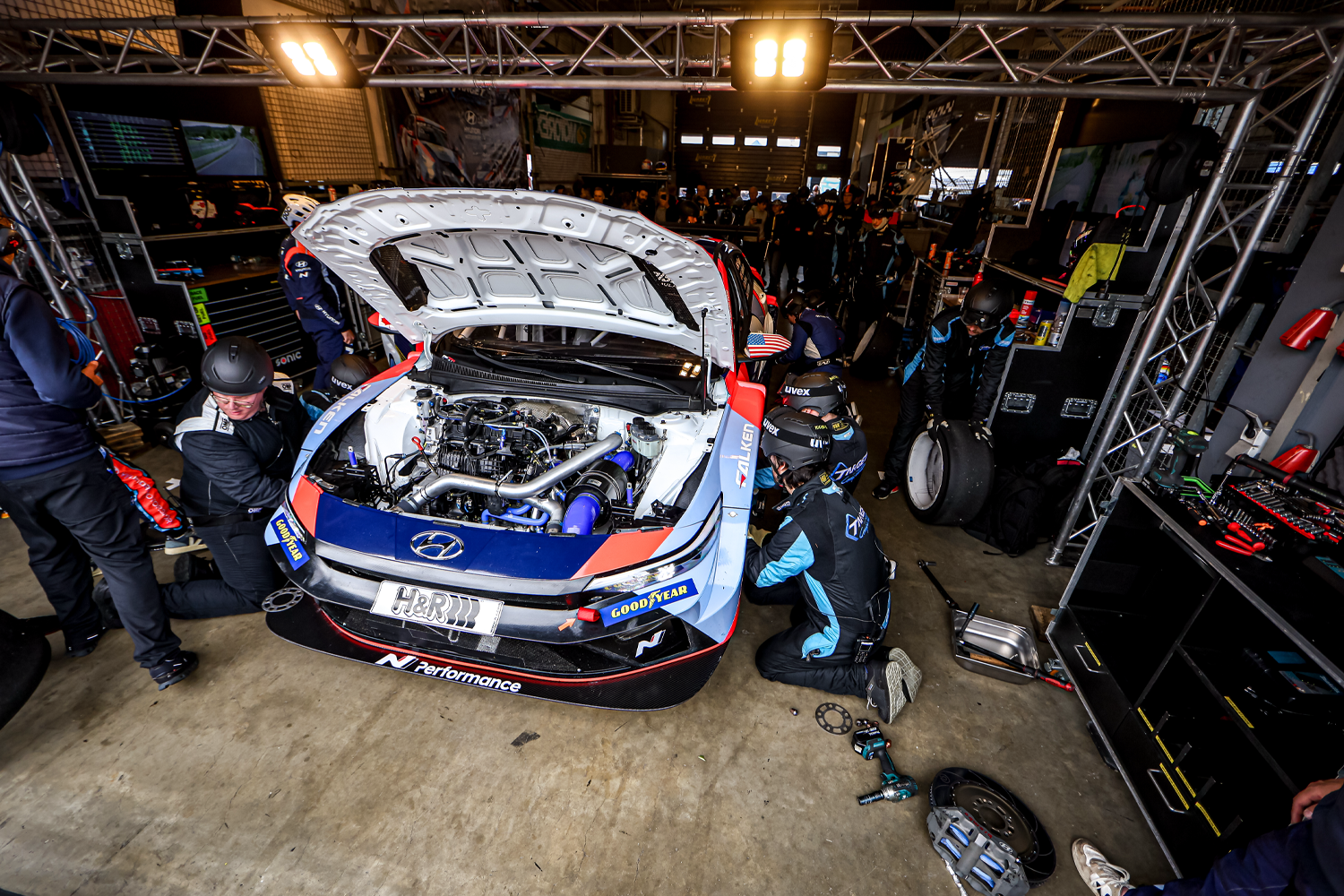 Hyundai N Performance race car in a pit garage with the hood open, as a team of mechanics works on various parts of the vehicle under bright lights, surrounded by tools and equipment.
