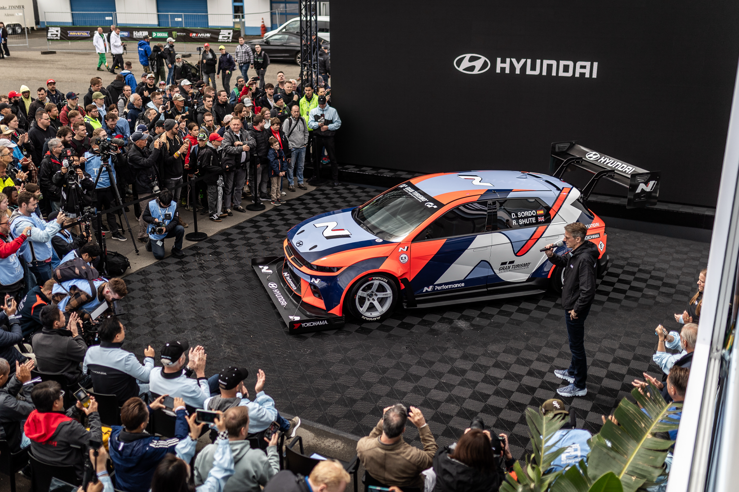 Hyundai N Performance race car displayed on a podium at a public event, surrounded by an audience, photographers, and media, with a presenter speaking about the car's features.
