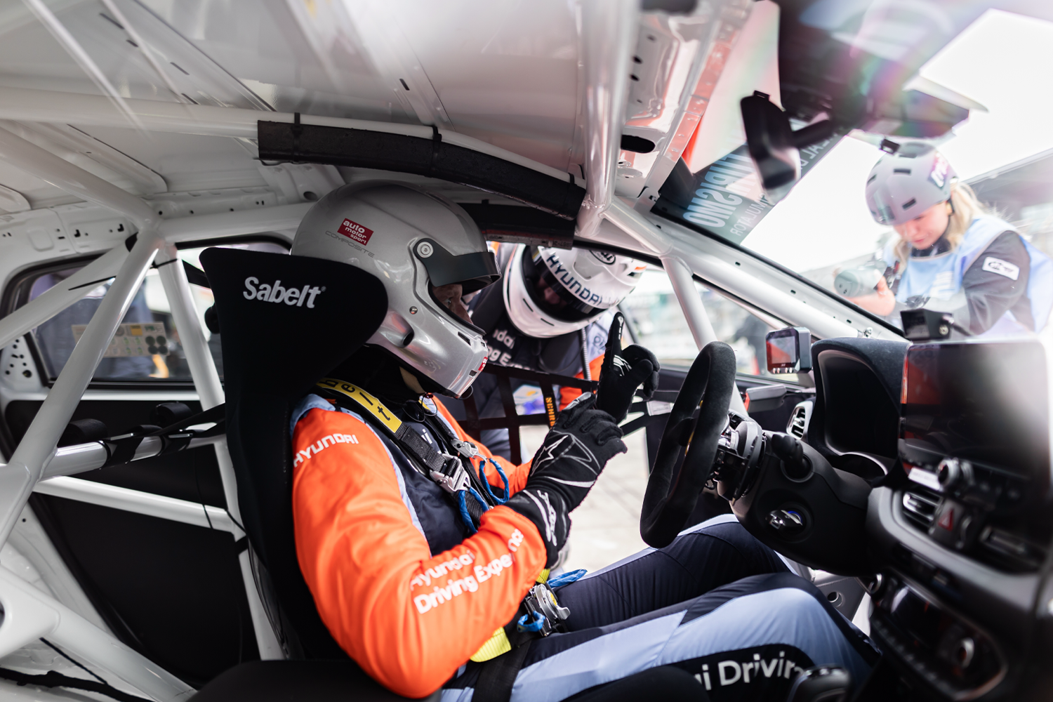 Professional racing driver seated in a Hyundai race car cockpit, wearing a helmet and full racing gear, engaging with a team member providing last-minute instructions outside the vehicle.
