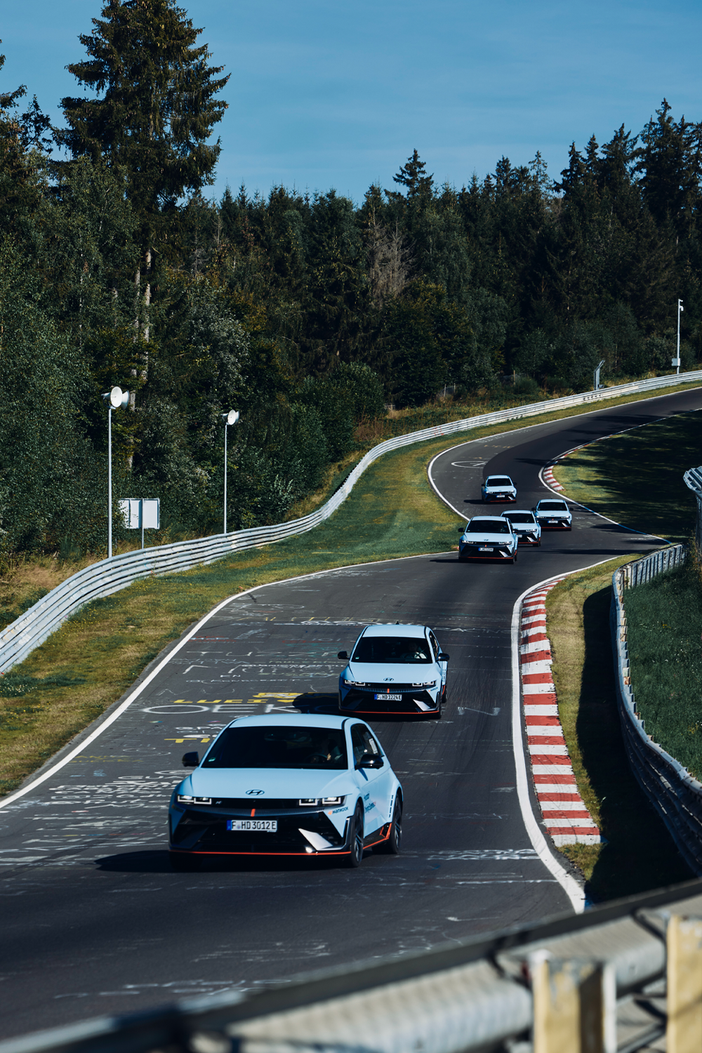 A convoy of Hyundai IONIQ 5 N cars driving on the Nürburgring Nordschleife, surrounded by lush greenery and graffiti-covered asphalt.
