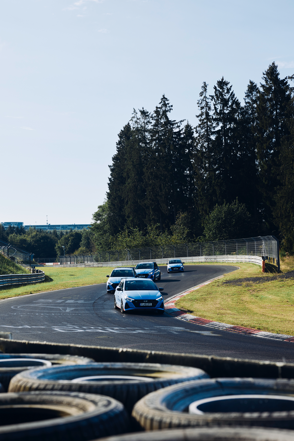 A group of Hyundai i20 N cars navigating a curve on the Nürburgring Nordschleife, with tire barriers in the foreground and tall trees in the background.
