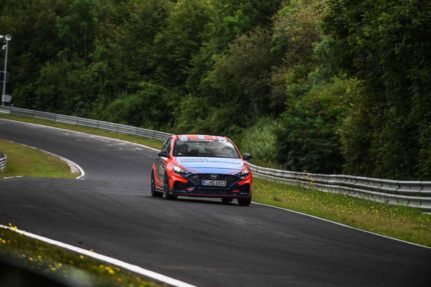 A Hyundai i30 N in Hyundai Driving Experience livery navigating a turn on the Nürburgring Nordschleife, surrounded by dense greenery.
