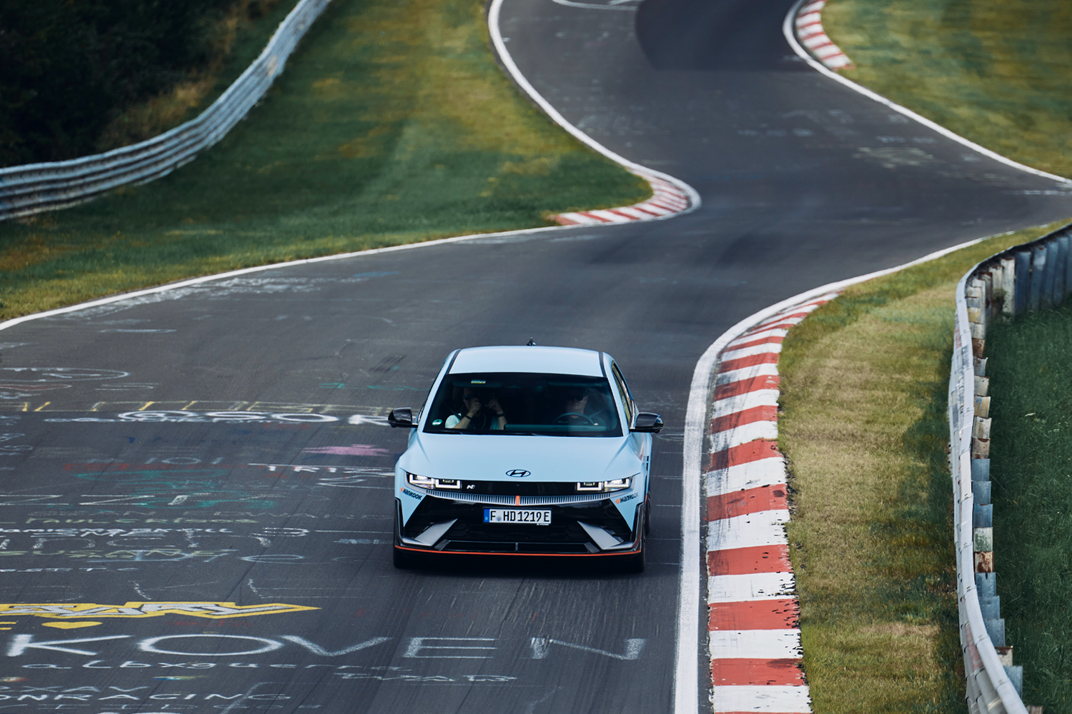 A Hyundai IONIQ 5 N driving on the Nürburgring Nordschleife, surrounded by graffiti-covered asphalt and winding curves.
