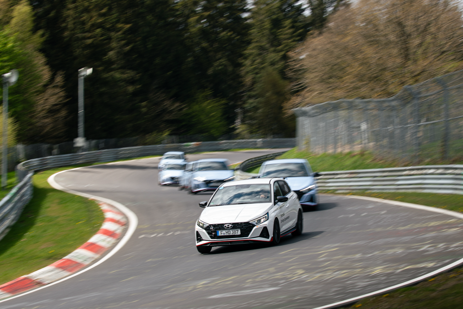 A fleet of Hyundai i20 N cars navigating a curve on the Nürburgring Nordschleife, with motion blur highlighting their speed and agility.
