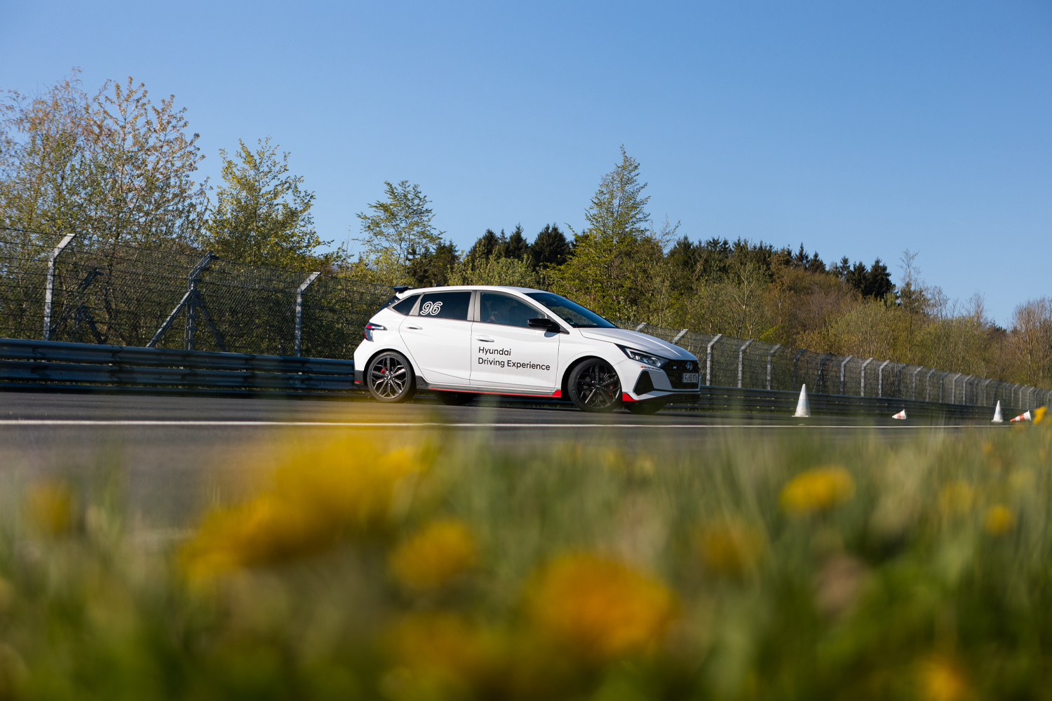A white Hyundai i20 N with Hyundai Driving Experience branding on the Nürburgring Nordschleife, framed by yellow wildflowers under a clear blue sky.
