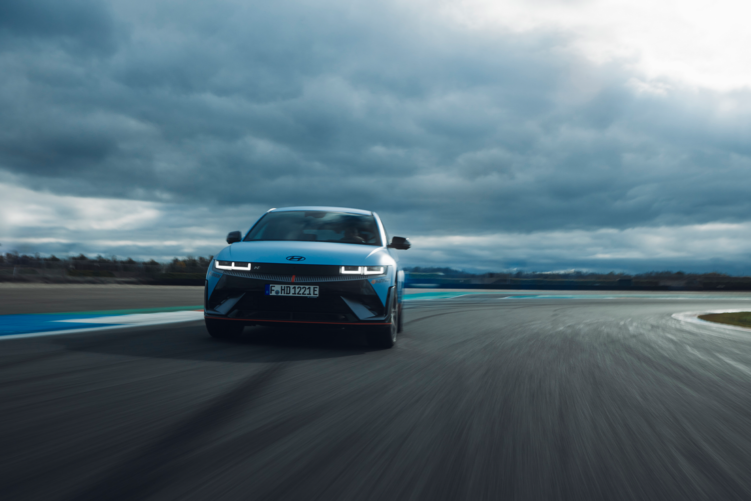 Hyundai IONIQ 5 N driving towards the camera on a racetrack with dramatic clouds overhead, highlighting its futuristic LED headlights and aggressive front design.
