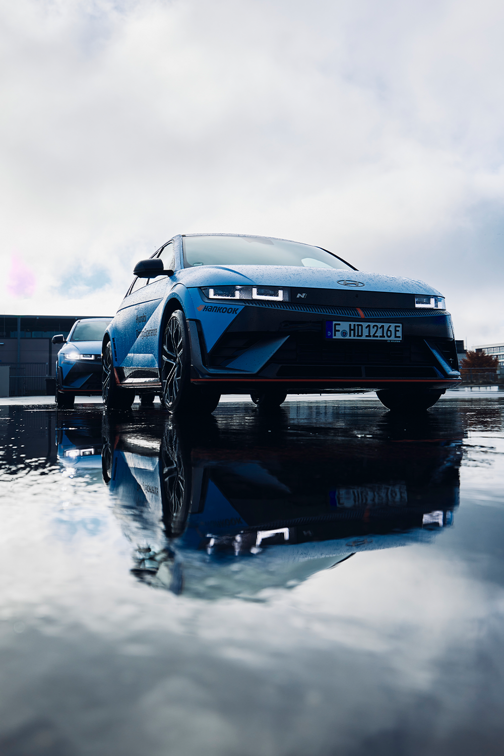 Hyundai IONIQ 5 N parked on a wet surface with its reflection clearly visible in a puddle, showcasing its aggressive front design and LED headlights under a cloudy sky.
