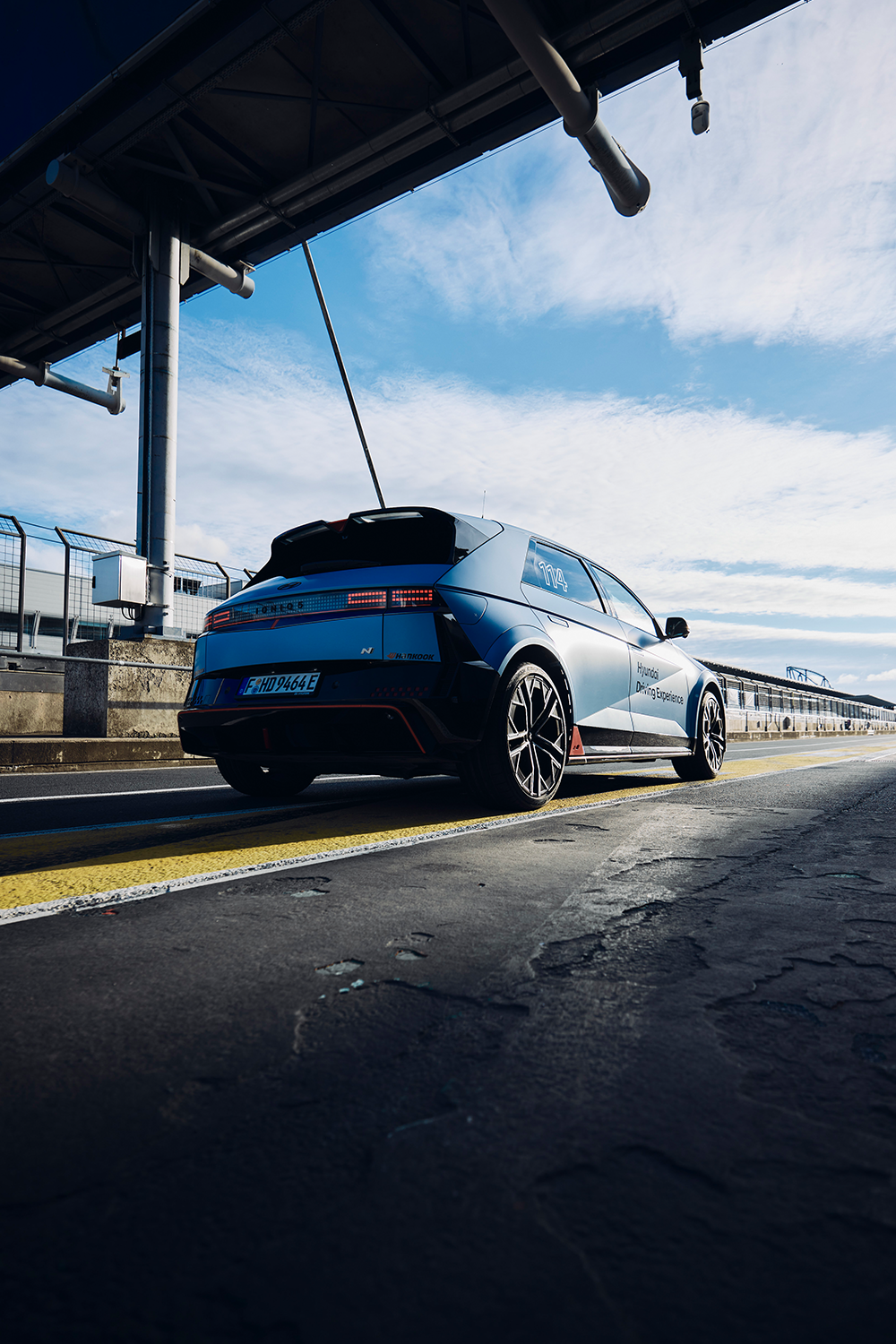 Rear three-quarter view of the Hyundai IONIQ 5 N parked in the pit lane under a structure, showcasing its sporty design, wide stance, and sharp tail lights against a clear blue sky.
