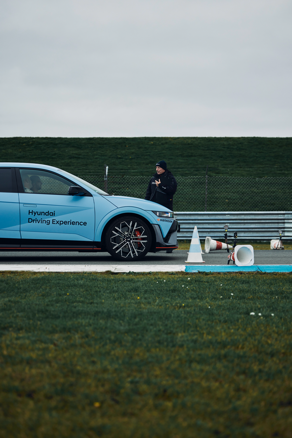 Hyundai IONIQ 5 N parked on a track during the Hyundai Driving Experience, with a crew member standing nearby and traffic cones scattered around.
