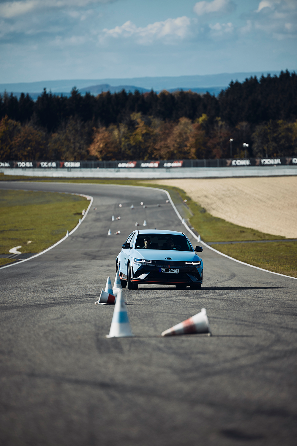 Hyundai IONIQ 5 N driving through a slalom course on a racetrack, maneuvering between traffic cones under a clear sky with a forested backdrop.
