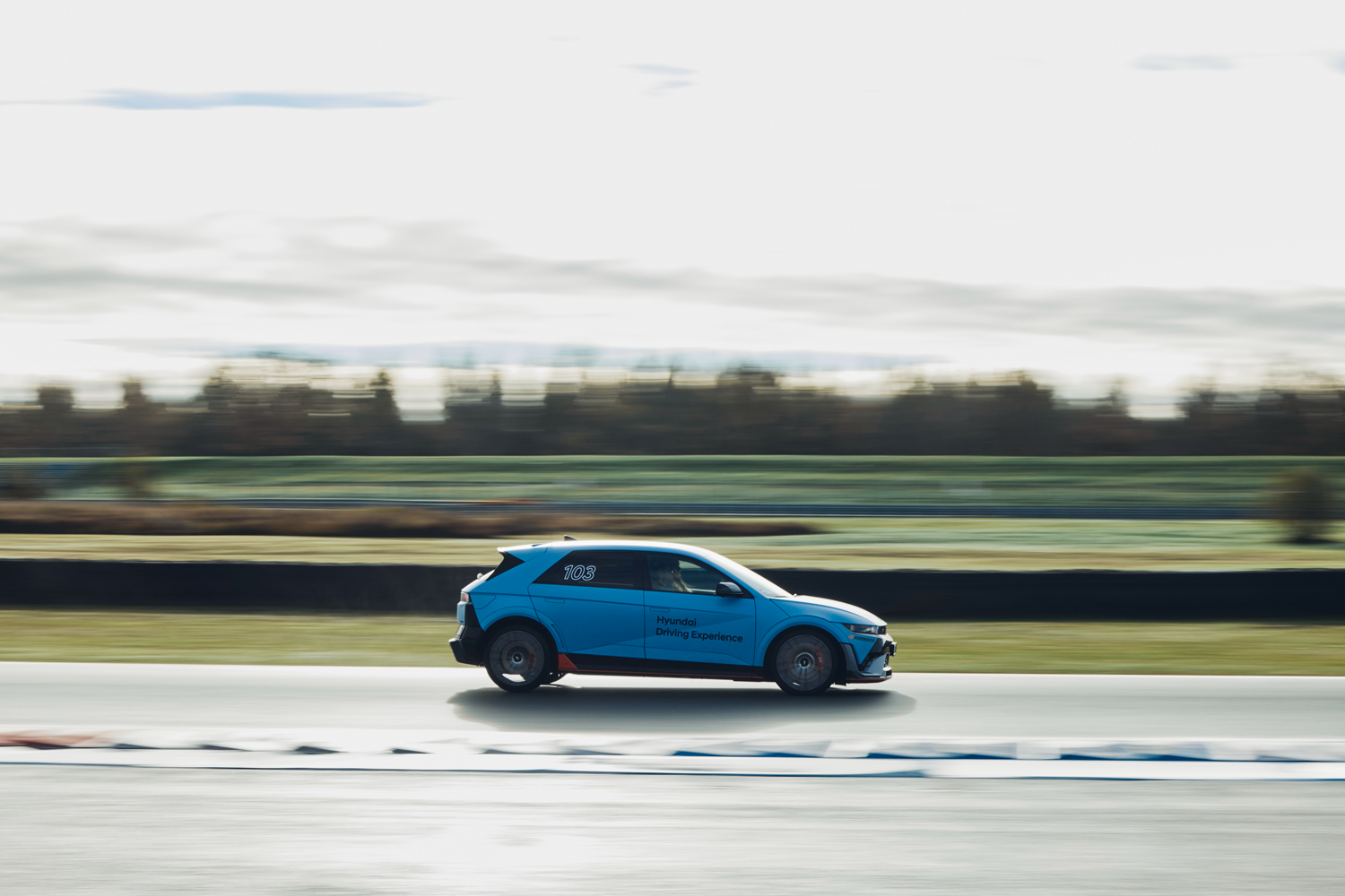 Side view of a Hyundai IONIQ 5 N driving on a racetrack with motion blur and featuring the 'Hyundai Driving Experience' branding.
