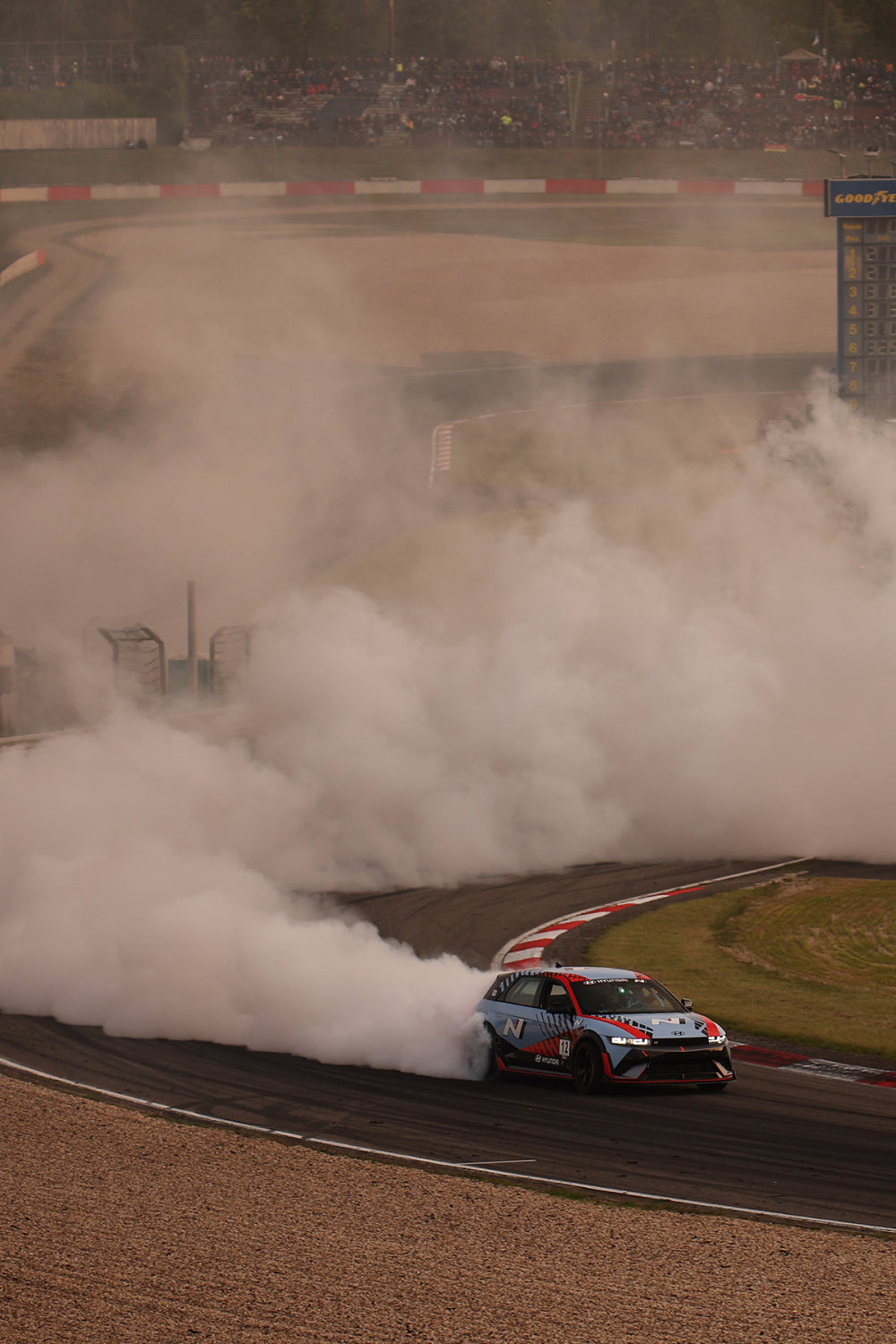 Hyundai N racing car drifting through a turn on a racetrack, producing thick clouds of smoke as spectators watch from the stands in the background.
