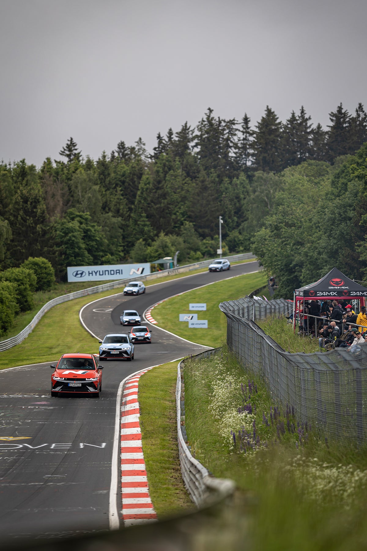 A lineup of Hyundai N vehicles driving along a curved section of the Nürburgring racetrack, surrounded by lush greenery, with spectators watching from a fenced area on the right.
