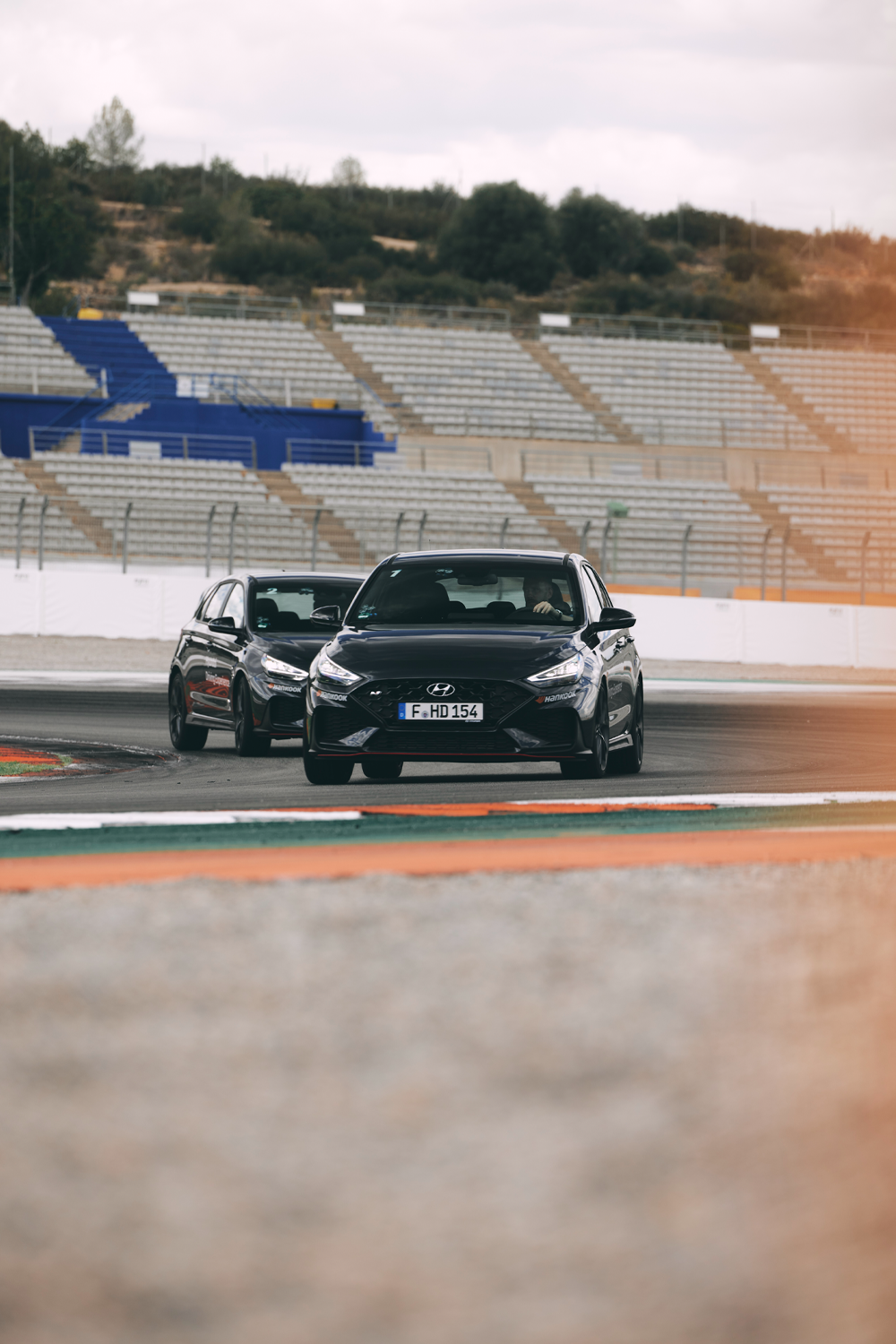 Two Hyundai i30N performance hatchbacks on a race track, with one black and one white car navigating a corner. The scene is set against empty spectator stands and an overcast sky.

