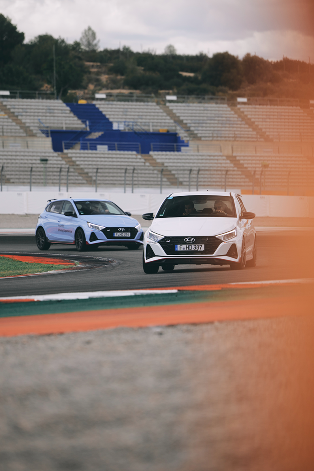 Two white Hyundai i20 N cars in action during a track driving session, navigating a sharp curve with precision. The foreground features blurred orange accents, adding a dynamic effect, while the background shows an empty grandstand under overcast skies.
