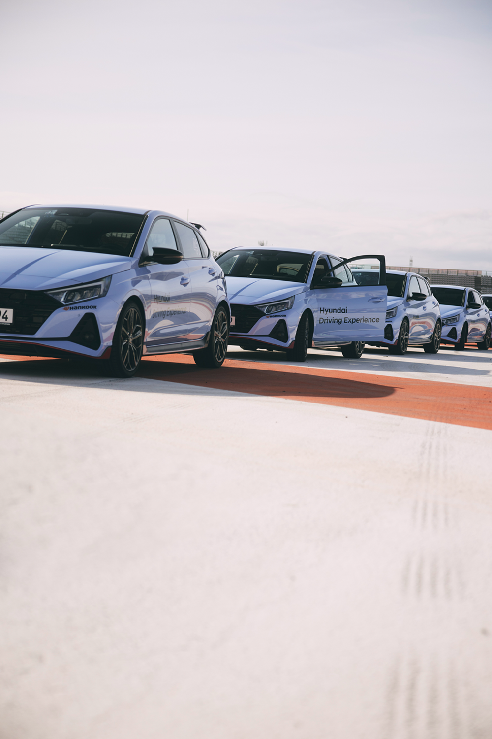 A line of Hyundai i20 N vehicles parked on a trackside, one with its door open, showcasing the Hyundai Driving Experience branding. The vehicles are light blue with sporty accents, positioned on a track marked with orange and white patterns, under a clear sky.
