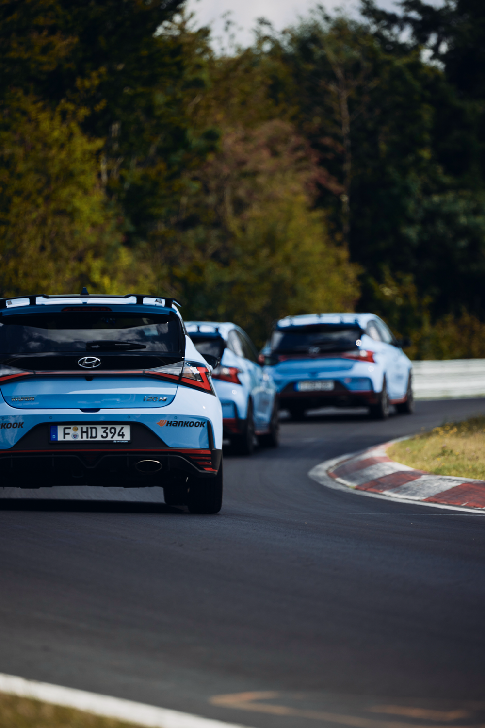 Three Hyundai i30 N cars in a convoy on a race track, captured from the rear perspective. The blue performance hatchbacks navigate a sharp curve, with trees and greenery in the background.
