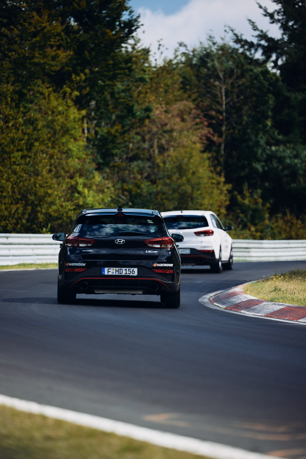 Two Hyundai i30 N vehicles on a race track, with one black car in the foreground and a white car following closely behind. The track curves through a green, wooded area with vibrant autumn foliage.
